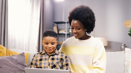 Portrait of happy African American family pretty mother and small son child sitting on sofa browsing online on laptop at home. Mom and little boy studying on computer, e-learning, education concept