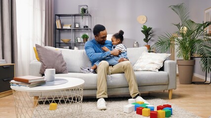 Cheerful young African American male parent playing with small adorable girl kid sitting on couch in modern living room. Dad tickling little daughter and laughing having fun a home together