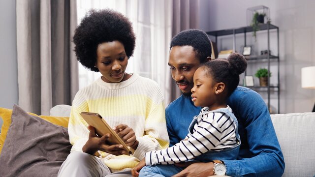 Portrait of cute African American family parents and little girl kid sitting on couch in living room at home and tapping on tablet teaching daughter using educative app on device, parenting concept