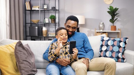 Portrait of African American happy joyful young male parent with small son waving hands while sitting on sofa in apartment and talking on online video call on smartphone. family time concept