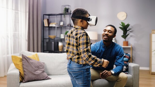 Little amazed African American boy teen wearing VR glasses standing in living room at home with young happy father, child in virtual reality headset using 3D futuristic technology, tech concept