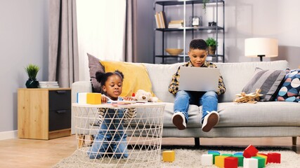 Two african american children spending weekends at home. Cute boy sitting on sofa and playing online games on laptop while little sister plays with colored markers.