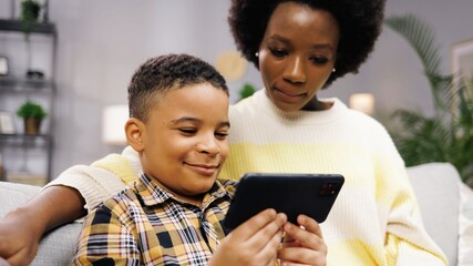 Portrait of cute happy African American little boy child typing on smartphone playing games while sitting on sofa with young mom parent at home. Using tech gadget, cellphone app, kid concept