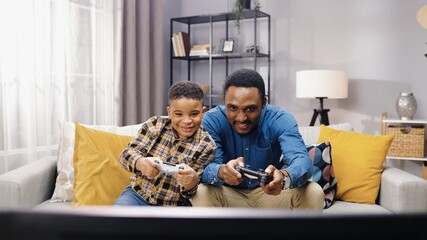 Portrait of African American joyful young male parent having fun with small son sitting on sofa in...