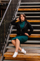 Fototapeta na wymiar Stylish young pretty hipster woman sitting on iron house stairs, Happy beautiful girl in casual clothes green dress, black jacket white sneakers, urban background, street photo, body positivity