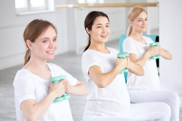 Fototapeta premium Happy young pregnant women working out with dumbbells on fitball in studio