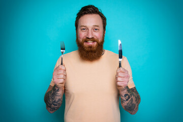 Man with tattoos is ready to eat with cutlery in hand. cyan background