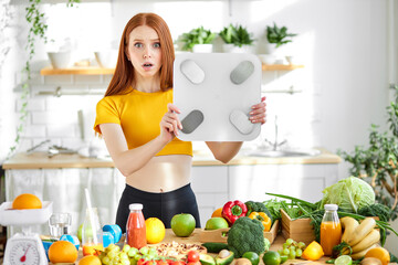 Redhead woman stand in shock holding weight scales, suprised by her weight, in kitchen