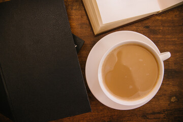 A white cup of coffee sits on a wooden table. Hardcover books lie nearby. Textbooks or fiction.