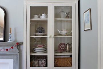 White wooden cupboard with clear glass used to store teapots, plates, tea cups and other kitchen accessories. 