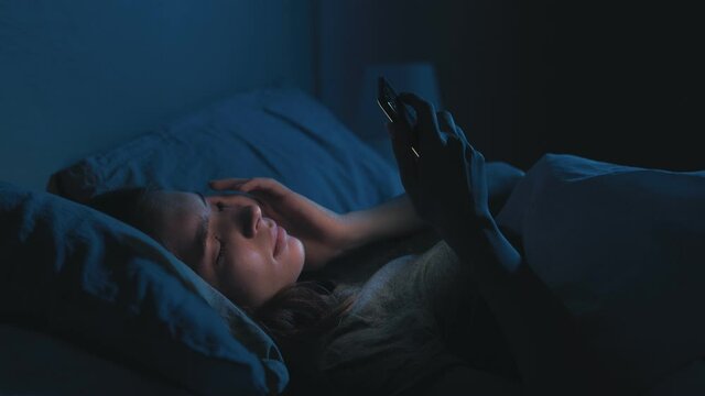 Late online. Night insomnia. Sleep disorder. Exhausted bored tired woman using mobile phone lying alone in bed in dark home bedroom with blue light.