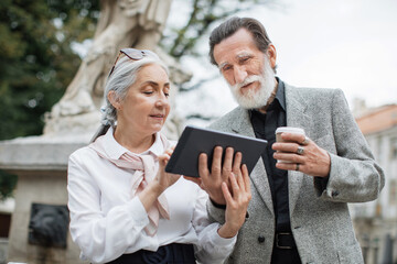 Bearded senior man holding digital tablet while his lovely wife pointing on screen. Two mature people sitting outdoors, drinking coffee and using modern gadget.