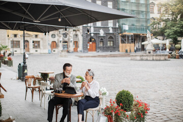 Happy mature family drinking coffee at cafe terrace. Stylish man and woman using tablet and retro camera. Relaxing outdoors after successful shopping.