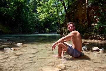 A handsome Caucasian man sitting on a rock in a river under sunlight