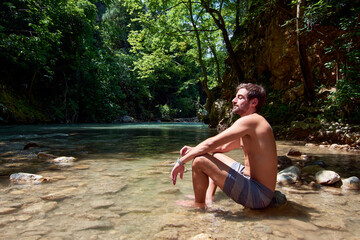 A handsome Caucasian man sitting on a rock in a river under sunlight