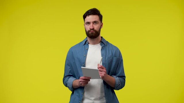 A portrait of a Caucasian who thinks about something and then writes something down in a notebook with a pencil. Guy in studio against yellow background. Close up. Slow motion ready 59.97fps.
