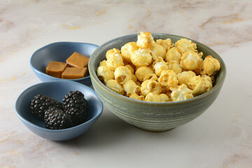 Caramel popcorn with a hint of blackberry liqueur in snack bowl and caramel squares and fresh blackberries in small ingredient bowls