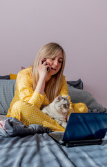 Young woman works on laptop sitting on bed in bedroom next to her pet