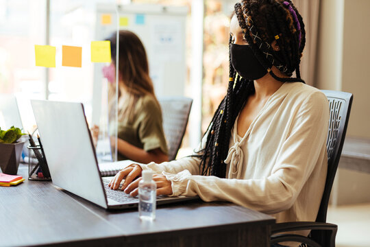 Business women wearing protective masks sitting at their desks separated by plexiglass dividers
