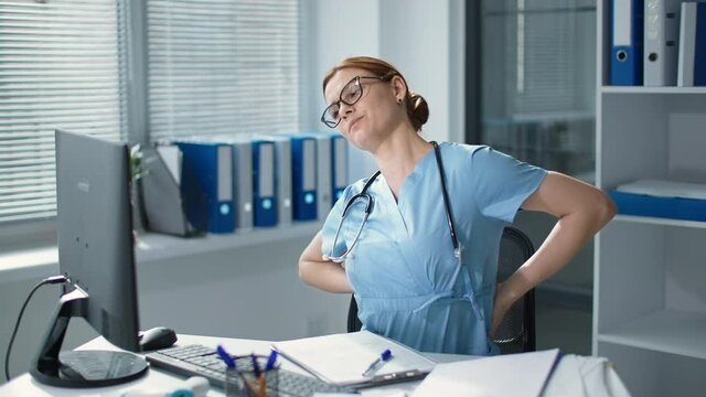 tired female doctor in glasses works at computer in hospital, sick young woman in medical uniform feels back pain sitting at the desk
