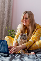 Young woman works on laptop sitting on bed in bedroom next to her pet