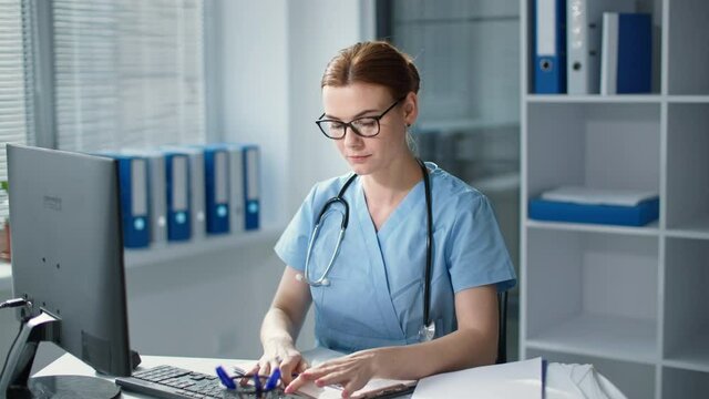 tired female doctor works at the computer and then taking off glasses sitting at desk, young woman in blue uniform with stethoscope feels headache in hospital