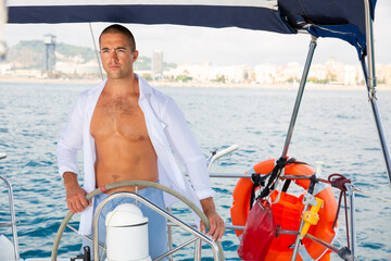 Handsome man steering modern yacht in Mediterranean sea along Barcelona coastline on sunny day..