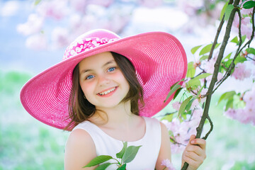 Beautiful small teen girl in pink fashionable hat with smiling face in spring flower blossom on sunny day. Spring teenager portrait.