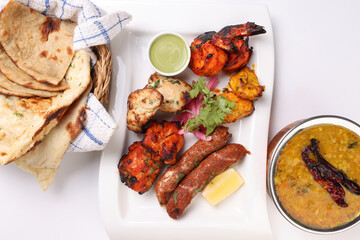 Mixed grill kebab chicken mutton beef prawn fish mint sauce lemon pickled onion bread basket chapati naan tandoori paratha dal lentil fry tadka copper pot on white dish over white background