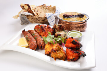 Mixed grill kebab chicken mutton beef prawn fish mint sauce lemon pickled onion bread basket chapati naan tandoori paratha dal lentil fry tadka copper pot on white dish over white background