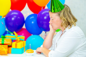 A middle-aged woman in a festive cap sits and sadly looks at a small cake with one candle. Fear of aging concept