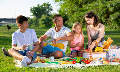 Cheerful family of four picnicking on green lawn in summer city park..
