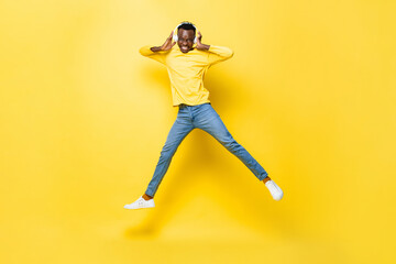 Fototapeta na wymiar Happy young African man wearing headphones listening to music and jumping in yellow isolated studio background