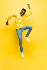 Fototapeta na wymiar Happy young African man wearing headphones listening to music and jumping with hand up in yellow isolated studio background