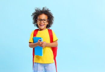 Cheerful little ethnic schoolchild with backpack and textbooks in studio - obrazy, fototapety, plakaty