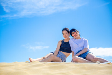 Fototapeta premium Two Chinese teenage girls leaning against each other in the sand dunes under sunny blue sky in holidays. 
