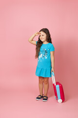 full-length portrait of a positive curly-haired girl carrying a suitcase and looking far away with hand over head, isolated on a pink background.