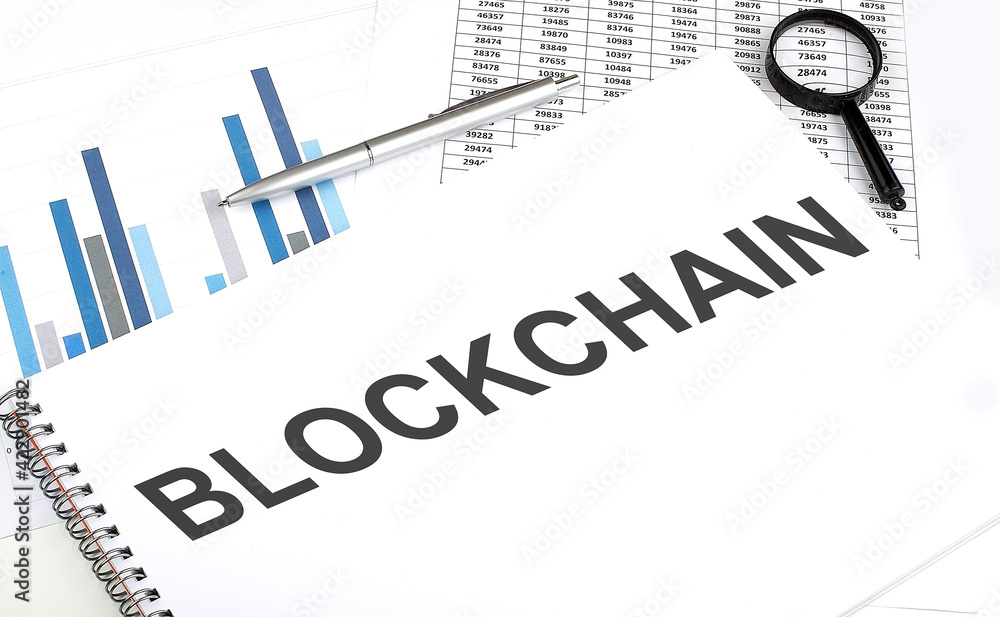 Poster in the notebook, the text blockchain . , next to magnifier and a pen, the background is graphics.