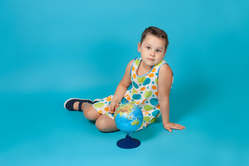 full-length portrait of a cute girl in a white summer dress sitting on the floor with a small globe, isolated on a blue background.