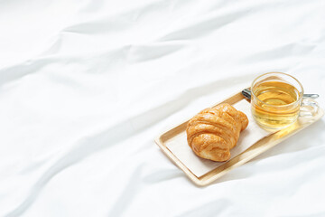 breakfast in bed concept with fresh croissant and hot herbal tea on brown wooden tray placed on white crumpled bedsheet.