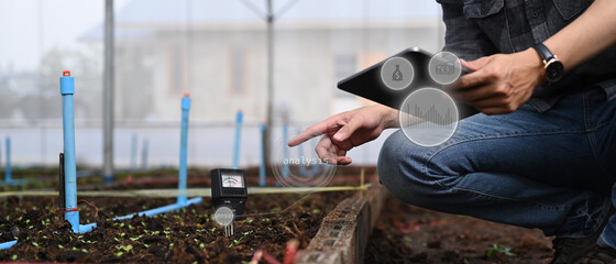 Close up view of agronomist using digital tablet for checking soil quality or pH value before seed plant in the greenhouse. Agriculture technology concept.