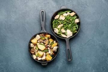 Frying pans with tasty vegetables and chicken on color background
