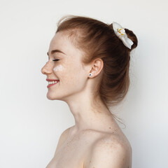 Adorable woman with red hair is smiling on a white studio wall holding a flower in hair posing with...