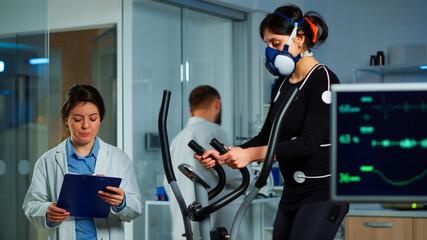 Specialists supervising exercises of patient, reading health information in clipboard, performance athlete running on cross trainer measuring heart rate, phisical endurance, psychological resistance
