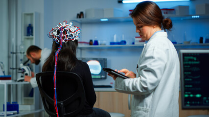 Obraz premium Back view of woman patient wearing performant eeg headset sitting on chair in neurological research laboratory while medical researcher adjusting it, examining nervous system typing on tablet.