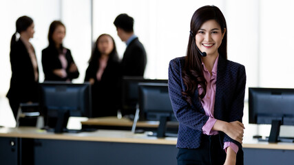 Young Asian woman in jacket and headset looking at camera with smile face and self confidence while working in call center with colleagues on blurred background. Service mind concept