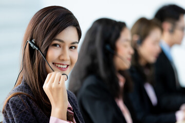 Cheerful Asian woman in headset smile with cheerful and happy while working on computer at desk with coworkers in help desk office. Sevice mind and ready for marketing support concept.