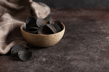Stack of of black potato chips in ceramic bowl on concrete table with copy space. Unusual chips. Unhealthy eat. Junk food.