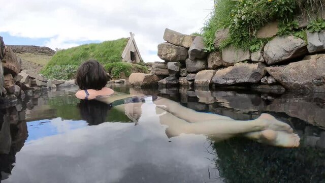 Woman relaxing in relaxing in Hrunalaug natural hot spring, geothermal hot pot water, Iceland. Traditional Icelandic turf house in the background 