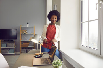 Young successful african american businesswoman in office portrait. Smiling black woman entrepreneur standing next to window rest after hard work looking at camera. Self-confidence and success